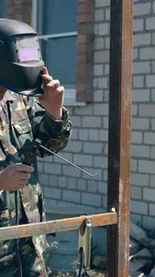 Trabajador suelda la futura carcasa de una cerca cerca de una casa de campo de ladrillo