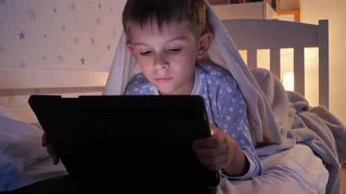 Portrait of cute boy in pajamas lying in bed and browsing internet on tablet computer