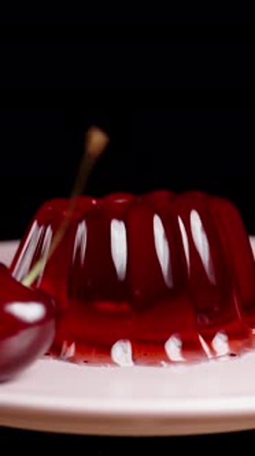 Vertical video. A cherry falls onto the red jelly on a pink plate, against a black background