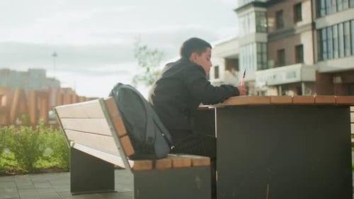 Side View Of Student Writing Outdoors At Bench Table Near School
