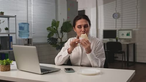 Geschäftsfrau sitzt am Schreibtisch im Büro und macht Mittagspause mit Essenslieferung, die am Laptop arbeitet