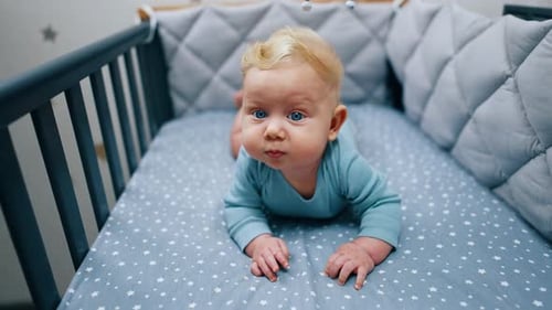 Cute baby boy lies in his crib. Blue-eyed blond child looking around. Close up.
