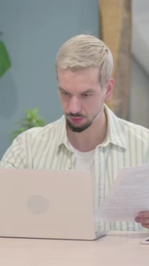 Young Man Looks at Documents with Laptop