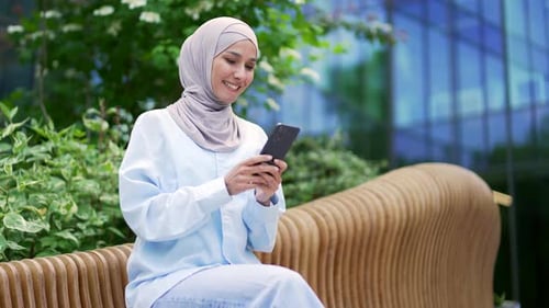 Young smiling muslim female employee in hijab is using a smartphone sitting on a bench on the street