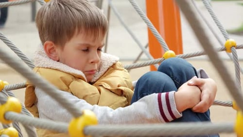 Upset and sad little boy being victim of bullying sitting alone on school playground