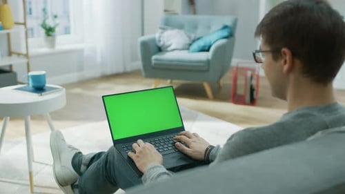 Over the Shoulder Shot: Young Man at Home Works on a Laptop Computer with Green Mock-up Screen. He'