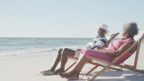 Happy senior african american couple on deck chairs and talking at beach, copy space, in slow motion