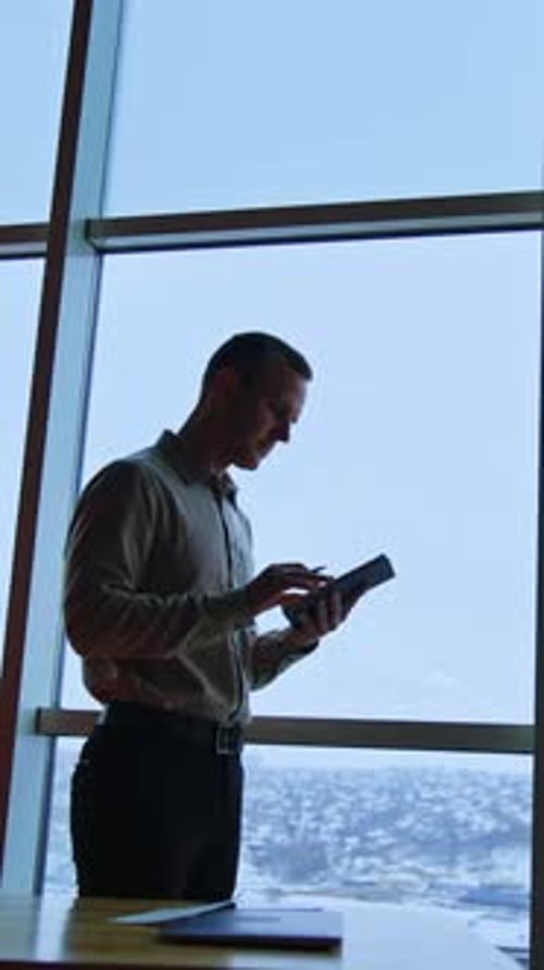 Handsome slim man stands at the window looking into his notebook. Office worker takes some notes.