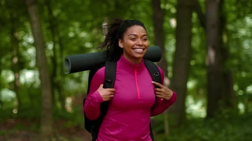Woman With Dark Skin Hiking Through a Green Forest While Carrying a Yoga Mat on Her Back