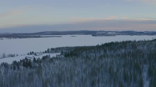 Aerial view across snow covered Scandinavian rural cabins surrounded by mountain range woodland wild