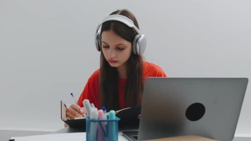 A Teenage Girl is Diligently Taking Notes in Her Notebook While Wearing Headphones and Looking at
