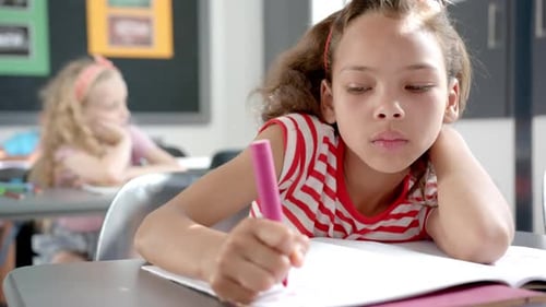 Biracial girl with light brown hair is focused on writing in a notebook in a classroom at school