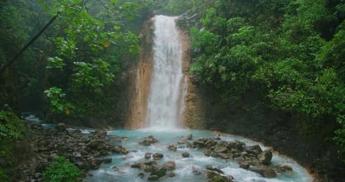 A tall waterfall pours into a rocky blue river in Costa Rica framed by dense rainforest during rainy