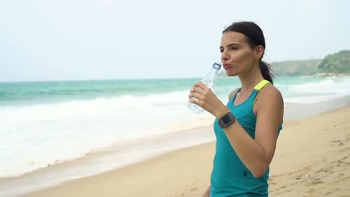 Sporty young woman drinks water after workout on exotic beach in super slow motion