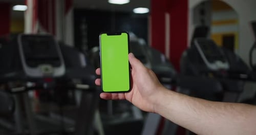 Man's Hand Holds Smartphone with Green Screen on Background of Gym Using Sports App