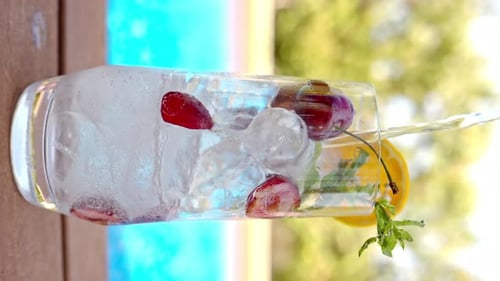 Mojito cocktail glass with ice and cherry and mint, soda and gin near a blue pool close up