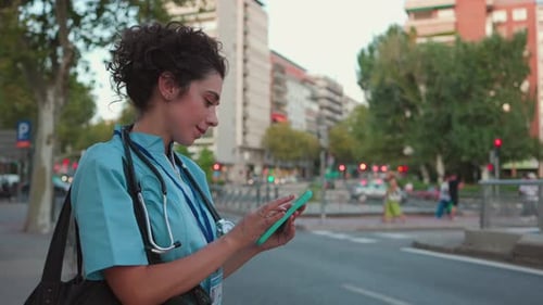 A young nurse in scrubs checks her phone while standing on a busy city street, focused and connected