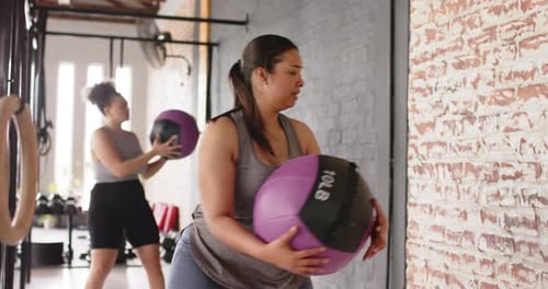 Diverse female duo twisting, throwing and catching medicine balls off wall in gym for toning core