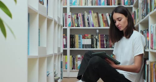 Young Girl Sitting on the Floor Near Shelves with Book in the Library Uses Digital Tablet Computer