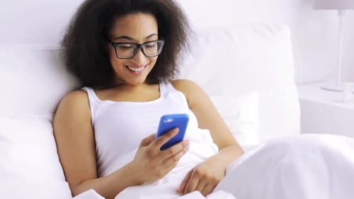 Young african american woman texts on smartphone relaxing in bed at home