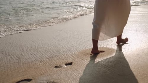 Barefoot Person in Flowing Dress Walks Along Sandy Shoreline Waves Gently Wash Over Footprints