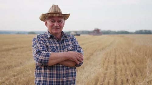 Happy Adult Farmer Stands and Smile at the Camera Against Background of Working Tractor in Field