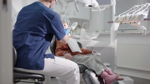 Friendly male dentist talks to smiling gap-toothed boy in chair after oral exam