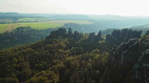 Rugged rock formations covered with trees, extending over green fields and distant hills. Schrammste