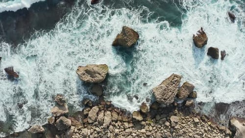 Aerial top down view of ocean waves crashing into golden rocky shore. Waves breaking into white foam