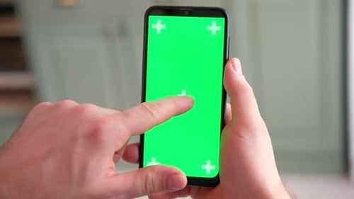 Young Man Sitting at Home Holding Smartphone Green Mockup Screen in Hand Male Person Using Chroma