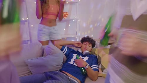 Sleepy young adult man lying down on sofa while his friends enjoy party.