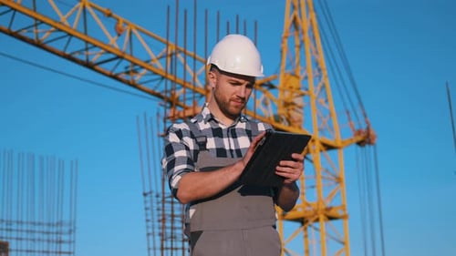 Construction worker that is uniform and hard hat is standing and using tablet