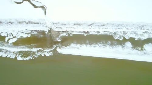 Vue aérienne de haut en bas d'un rivage hivernal glacé avec une plage enneigée et une eau de mer verte