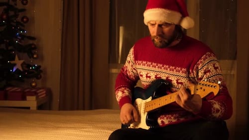 Young Man in Santa Hat Sitting and Playing Guitar Near Christmas Tree in Cozy Room Portrait Bearded