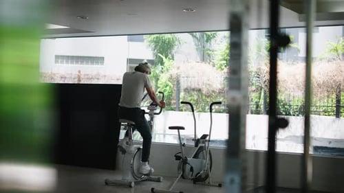Fitness Enthusiast Pedaling on an Exercise Bike in a Large Urban Gym Overlooking a Garden
