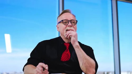 Adult Caucasian man in black clothes and red tie sits at desk.