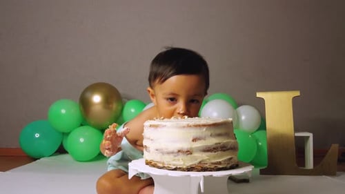 Cute latin baby toddler celebrating his 1st year birthday biting and smashing a cake with green ball