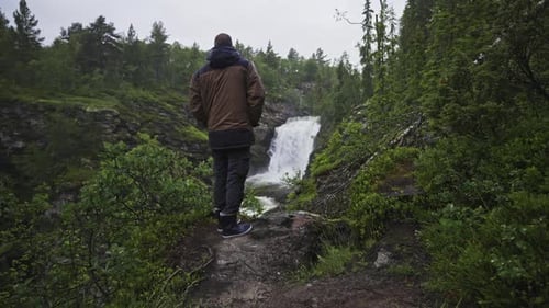 male enthusiast trekker walking in narrowed forest path reaching a scenic waterfall In Norway nation