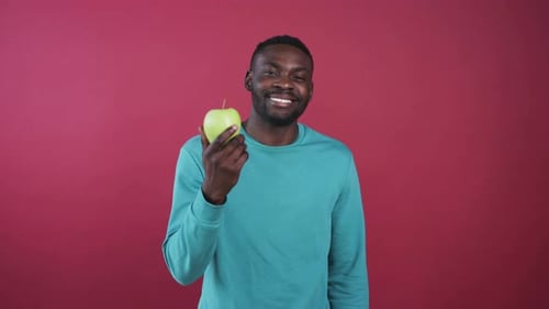 Likable African Guy with Shiny White Smile Holding Big Green Apple in Hand Looking at Camera Smiling