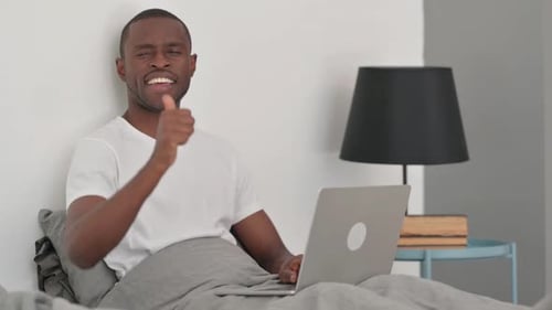 Homme africain avec les pouces levés regardant la caméra au lit avec un ordinateur portable