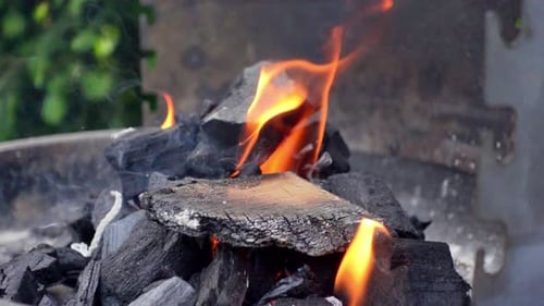 A slow-motion closeup shot focuses on coal igniting on fire with wood on a grill during a sunny day.