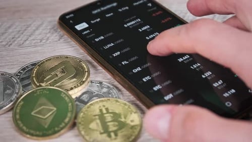 Trader Analyzes Cryptocurrency Prices on Smartphone on Table with Crypto Coins