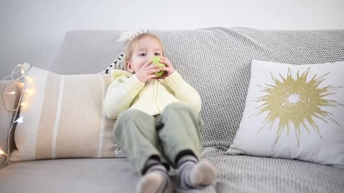 A Young Child Sits on a Cozy Sofa Enjoying a Green Apple with Curiosity and Contentment Surrounded