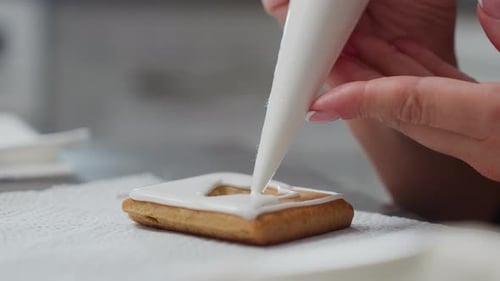 Closeup of Person Icing Cookie with White Icing From Piping Bag on Tissue Paper