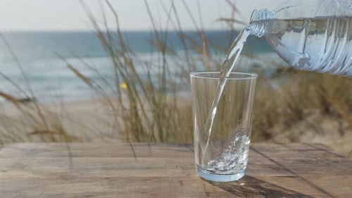 Wasser wird aus einer Plastikflasche in ein Glas gegossen, im Hintergrund Meerblick.