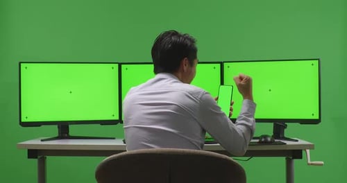 Happy Man Looking At Mobile Phone Green Screen And Celebrating While Working With Multiple Monitor