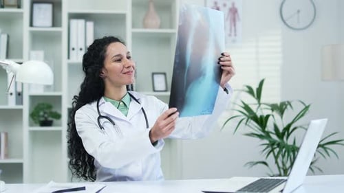 Confident female doctor in white coat examining x-ray picture while sitting at workplace in office i