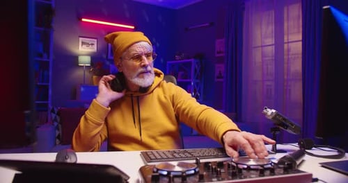 Close Up of Caucasian Stylish Senior Pensioner Male DJ Sitting at Table in Dark Room with Neon