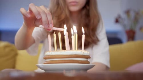 Close Up Pretty Young Brunette Lighting Candles on Birthday Cake in Room While Sitting on Yellow
