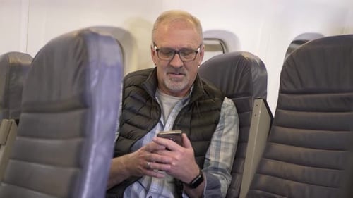 Senior man relaxes on airplane reading email and using smart phone
